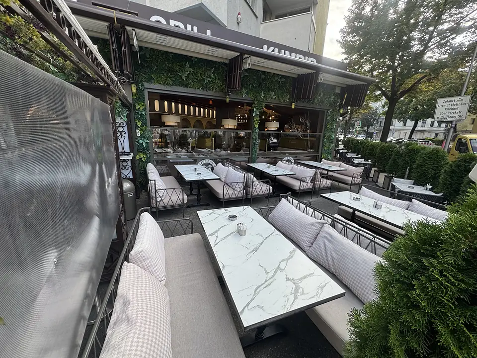 Überdachte Terrasse des Café Peri Berlin mit Polstermöbeln, Marmortischen und Heckenpflanzen als Sichtschutz.